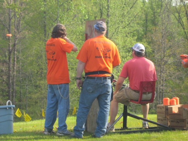 Male Shooter, Male Shotgun Coach, Thrower Puller, skeet in air to left of photo